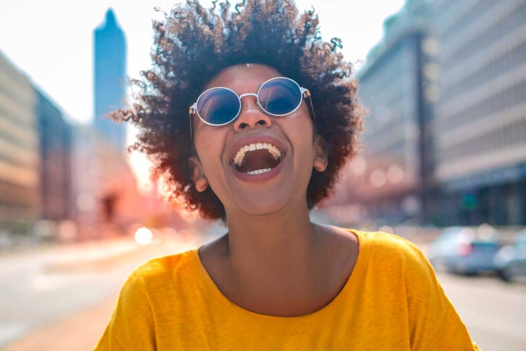 Plano coletivo por adesão: Foto de mulher negra com camisa amarela e óculos escuros rindo em uma avenida