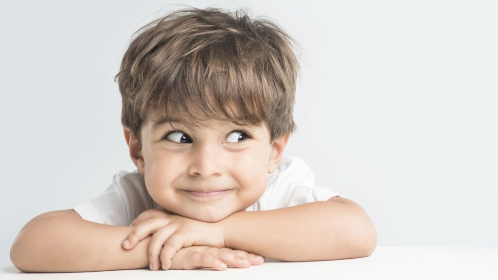 Plano de saúde para criança: foto de um menino sorrindo e apoiando sua cabeça sob suas mãos cruzadas em uma mesa